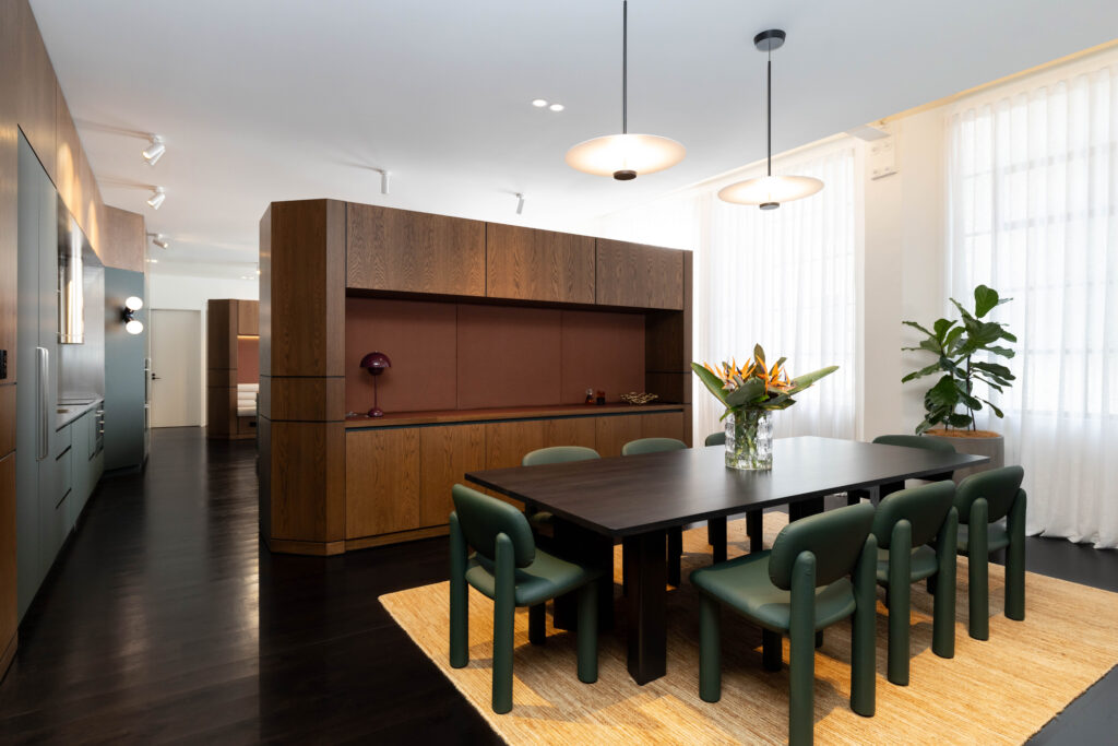 Modern office space with dark wood floors, a long black conference table with green chairs, a built-in wood cabinet, and large windows with white curtains.